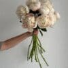 A hand holding a bouquet of light-colored peonies with long stems.